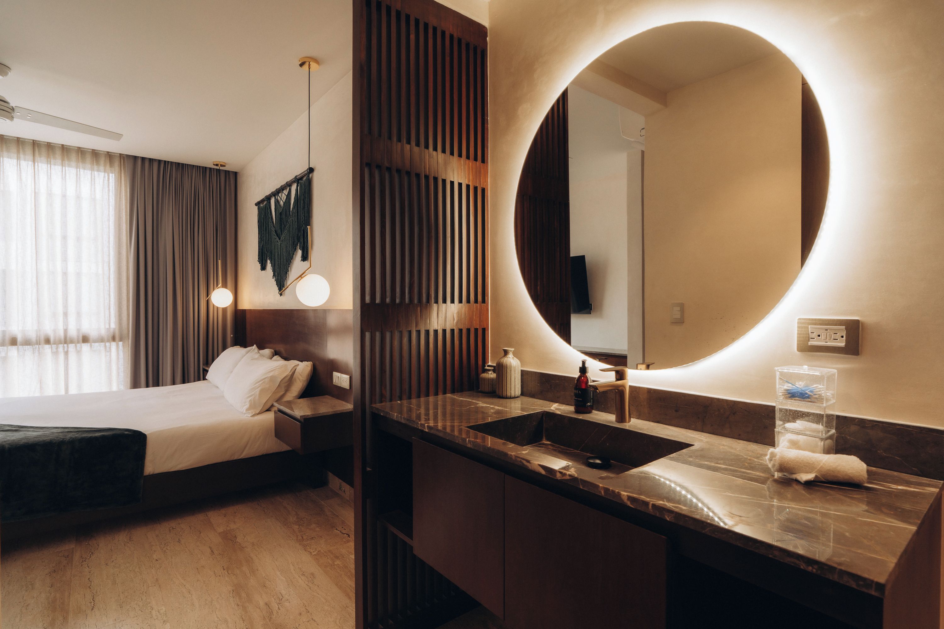 A hotel room with a bed, a round mirror, a sink, and a window with curtains.