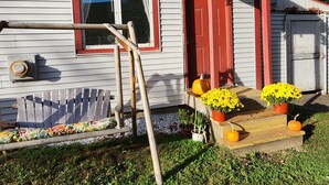 Una casa con puerta roja, ventana con molduras blancas, porche de madera con plantas en macetas y un banco de madera.