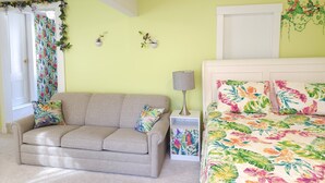 Un dormitorio con sofá, mesita de noche con lámpara, cama con ropa de cama floral y ventana con cortinas.