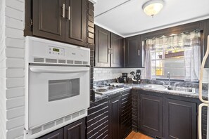A kitchen with dark cabinets, a white stove, and a window with sheer curtains. - Relax & Recharge, 2BR/1BA Lakefront Stay with Boat Slip + Pet-Friendly (Buchanan)