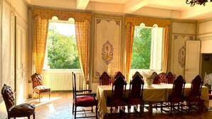Comedor con una mesa grande dispuesta para seis personas, rodeada de sillas ornamentadas. Hay dos ventanas con cortinas, un candelabro y un radiador.