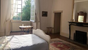 Un dormitorio con cama, una mesita, una silla, chimenea y ventana con cortinas.