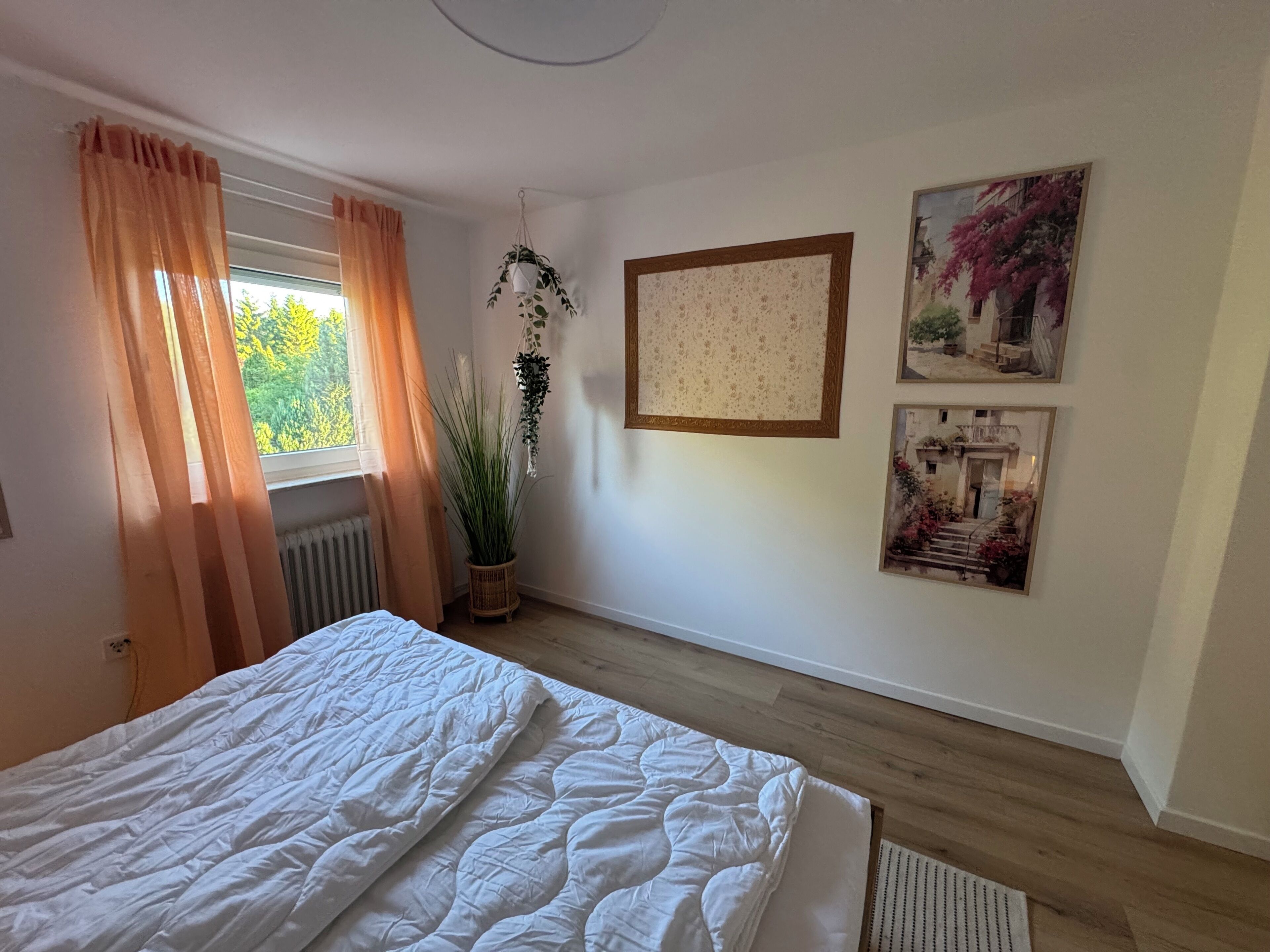 Ein Schlafzimmer mit Bett, Vorhängen an einem Fenster, einem Heizkörper, einer hängenden Pflanze und zwei gerahmten Bildern an der Wand.