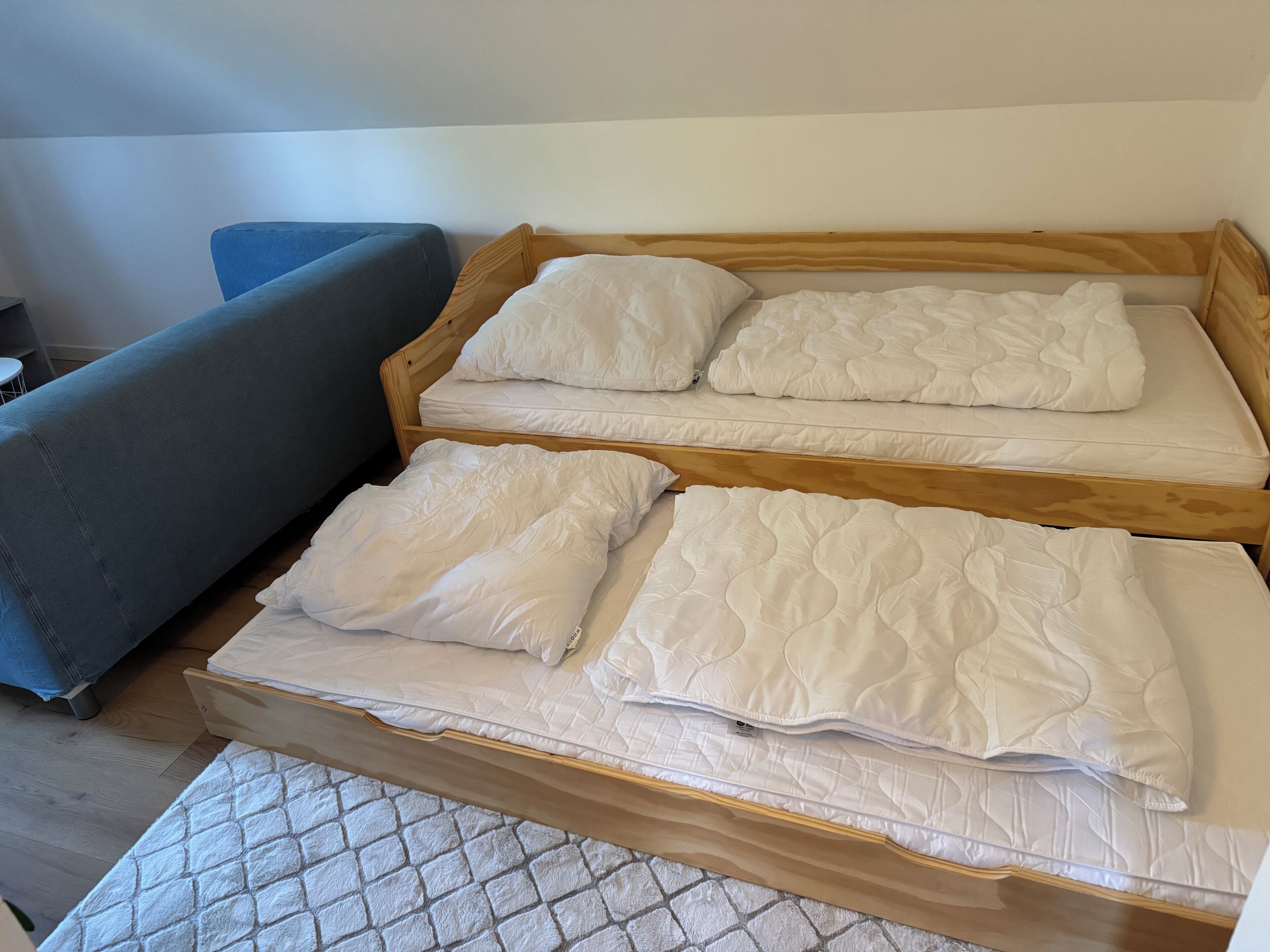 Una cama con ropa blanca y cabecera de madera en una habitación que incluye un sofá gris y una alfombra estampada.