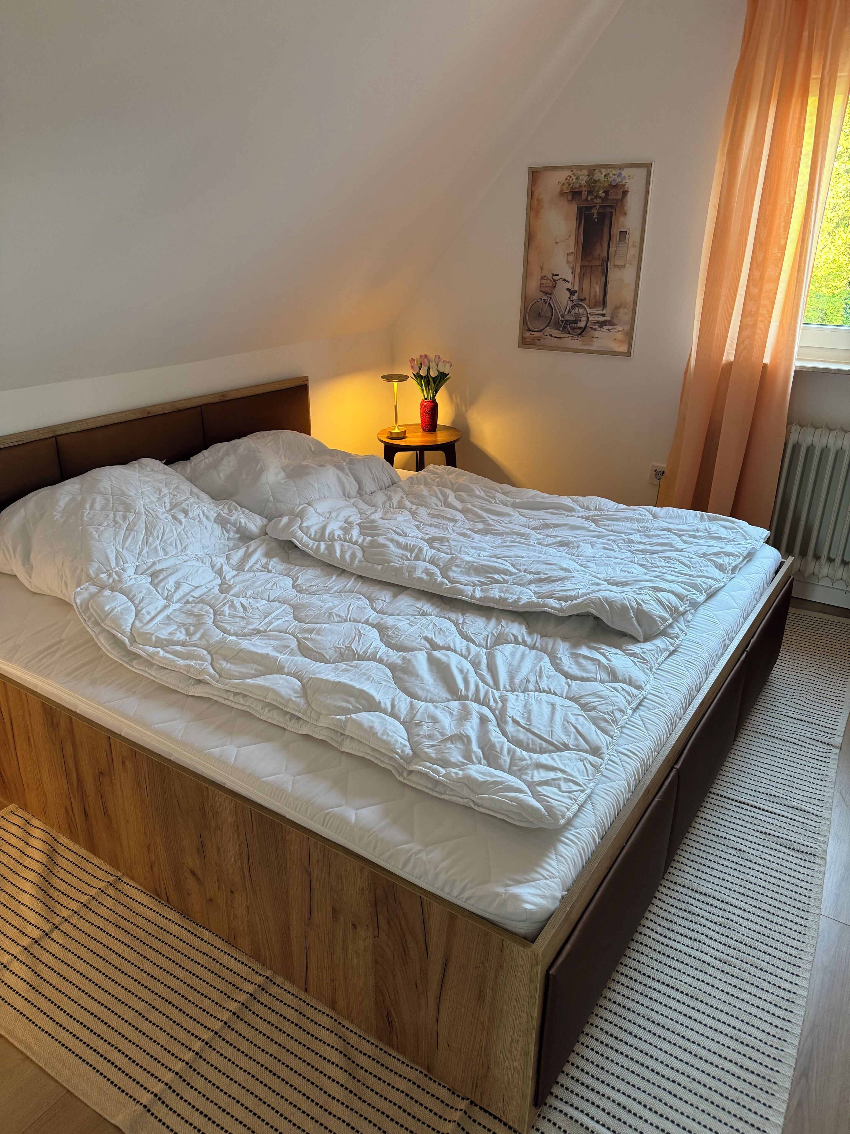 Ein ordentlich bezogenes Bett mit hölzernem Kopfteil, ein kleiner Tisch mit Blumen in einer Vase und ein Gemälde an der Wand.
