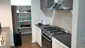 Eine moderne Küche mit Gasherd, Backofen und integrierter Dunstabzugshaube. Ein kleiner Arbeitsplatz mit Toaster und Mülleimer ist vorhanden. Die Küche verfügt über weiße Schränke und Holzboden.