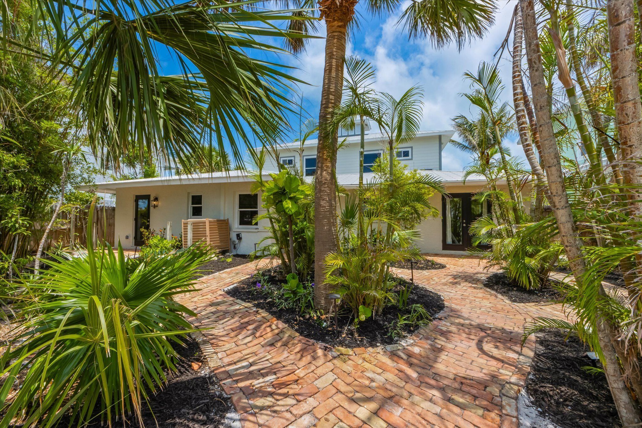 A modern house with a brick pathway, surrounded by palm trees and tropical plants.