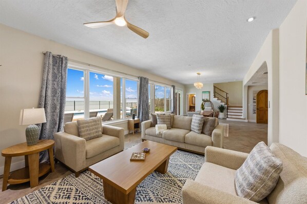 A spacious living room with a large window, a ceiling fan, and a staircase leading to an upper level. - Mimosa Bay: Waterfront Oasis w/ Heated Pool, Hot Tub & Bay Views (Anna Maria)
