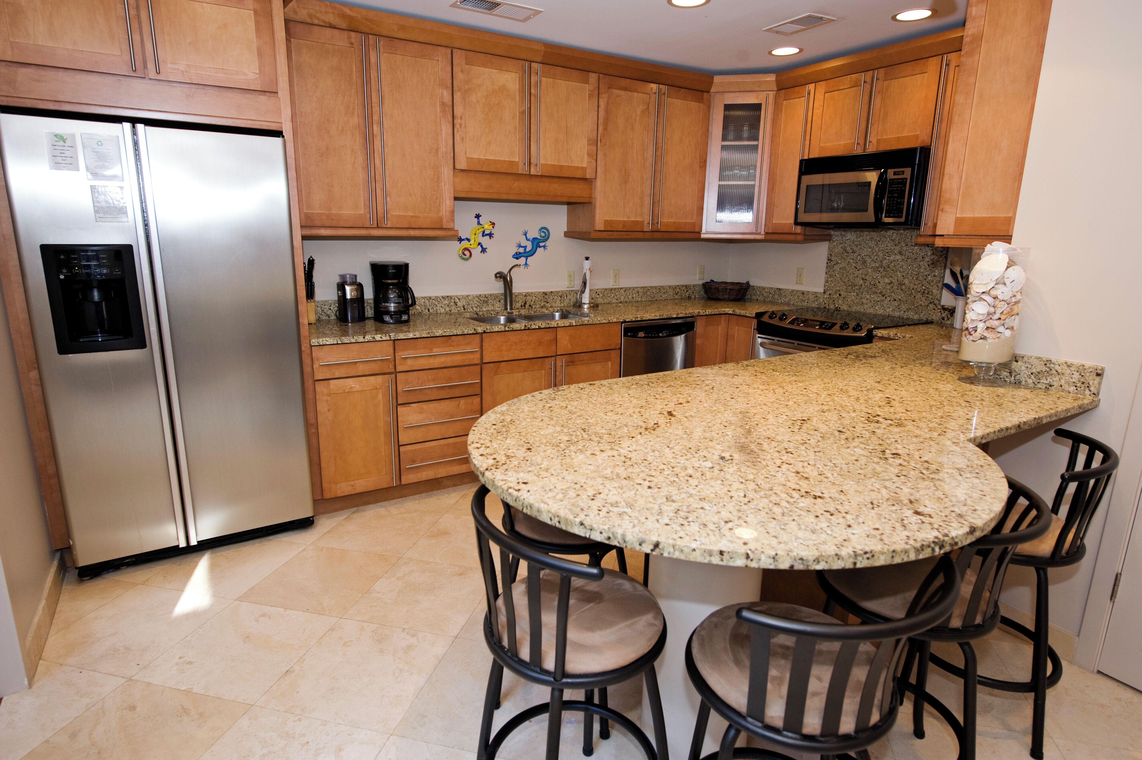 A kitchen with wooden cabinets, a stainless steel refrigerator, a granite countertop, a microwave, and a coffee maker.