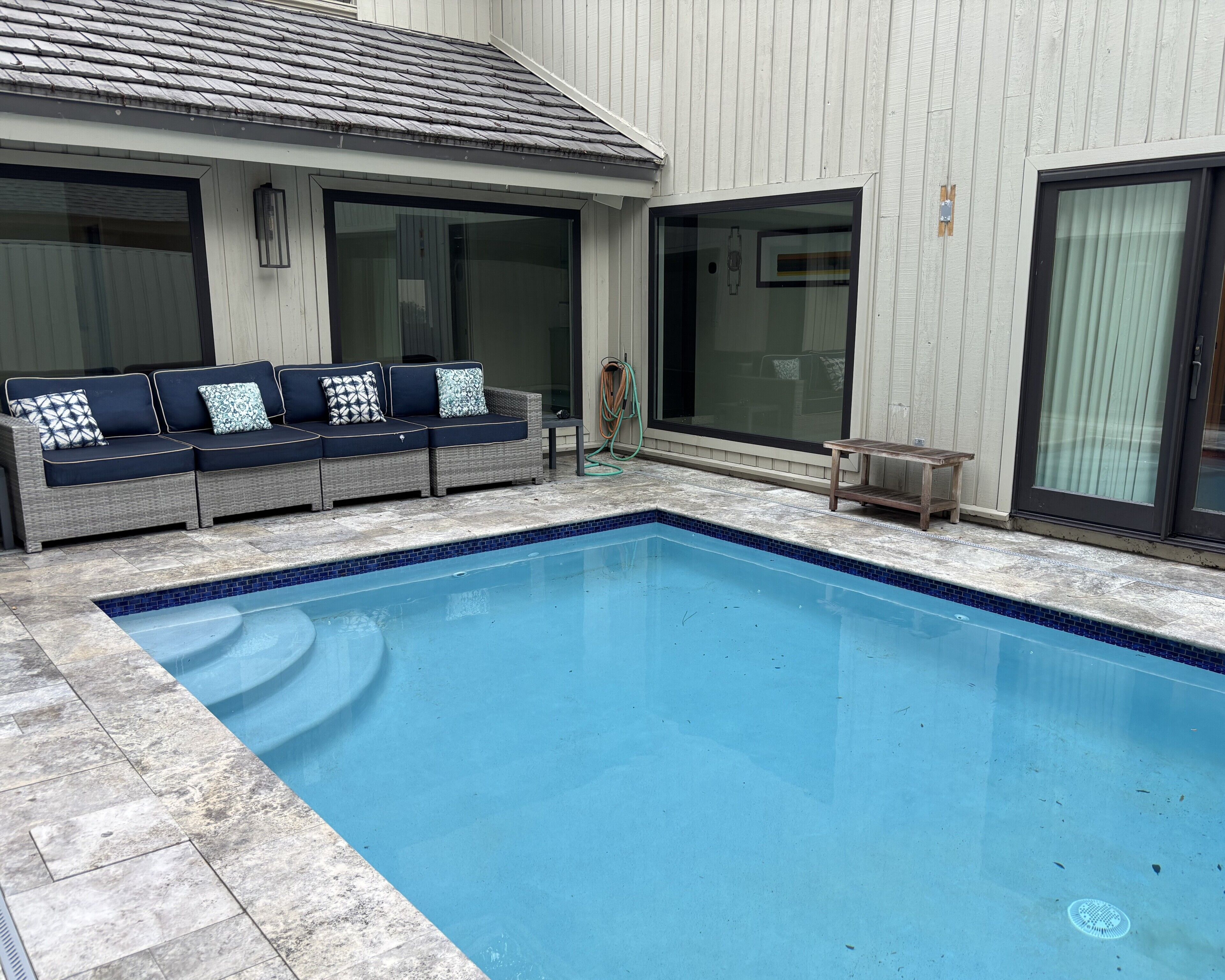 A swimming pool with a tiled edge, a gray outdoor sofa with patterned cushions, and a wooden table.