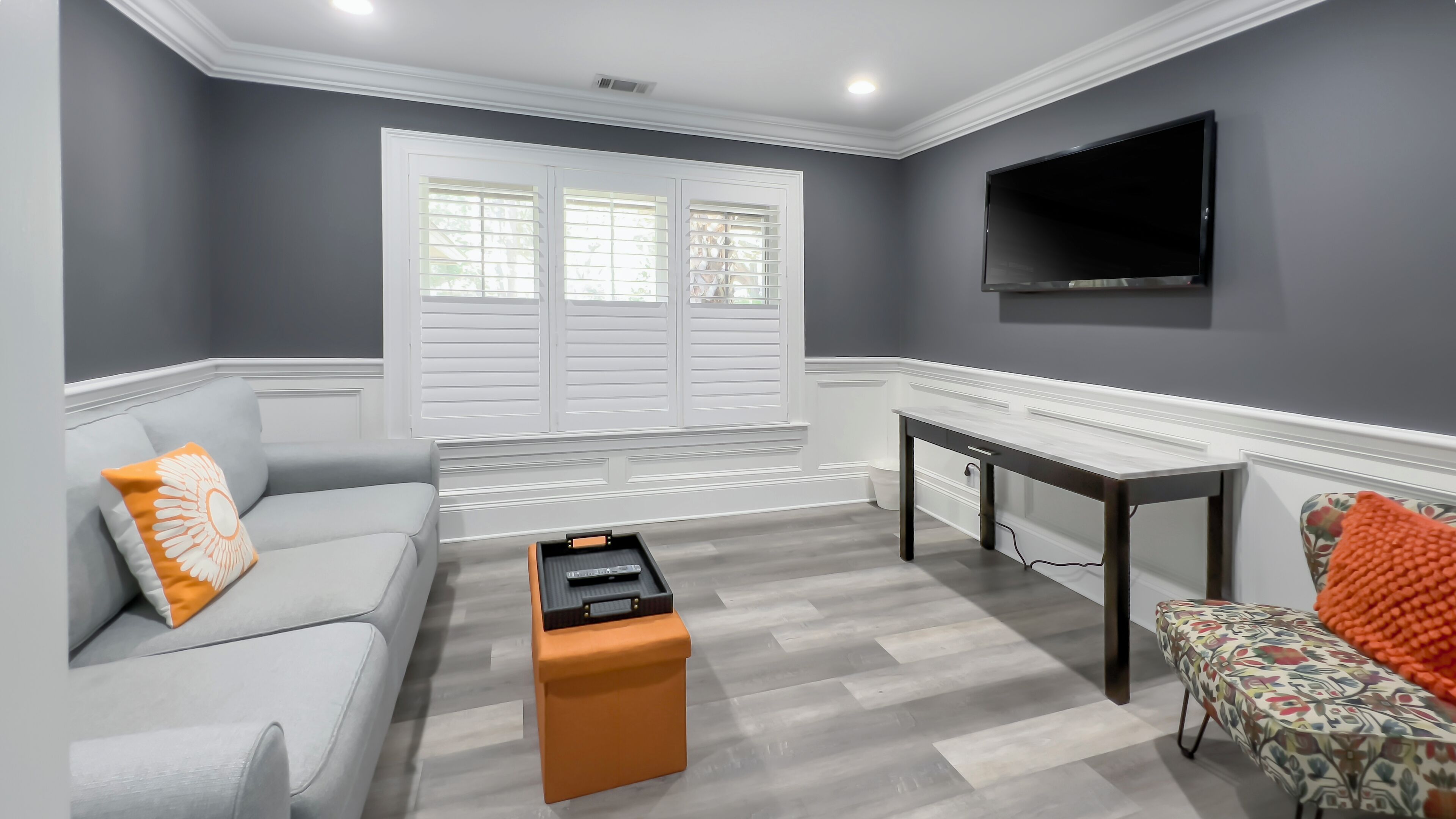 A living room with a grey sofa, a coffee table, a patterned armchair, a TV mounted on the wall, and a window with shutters.