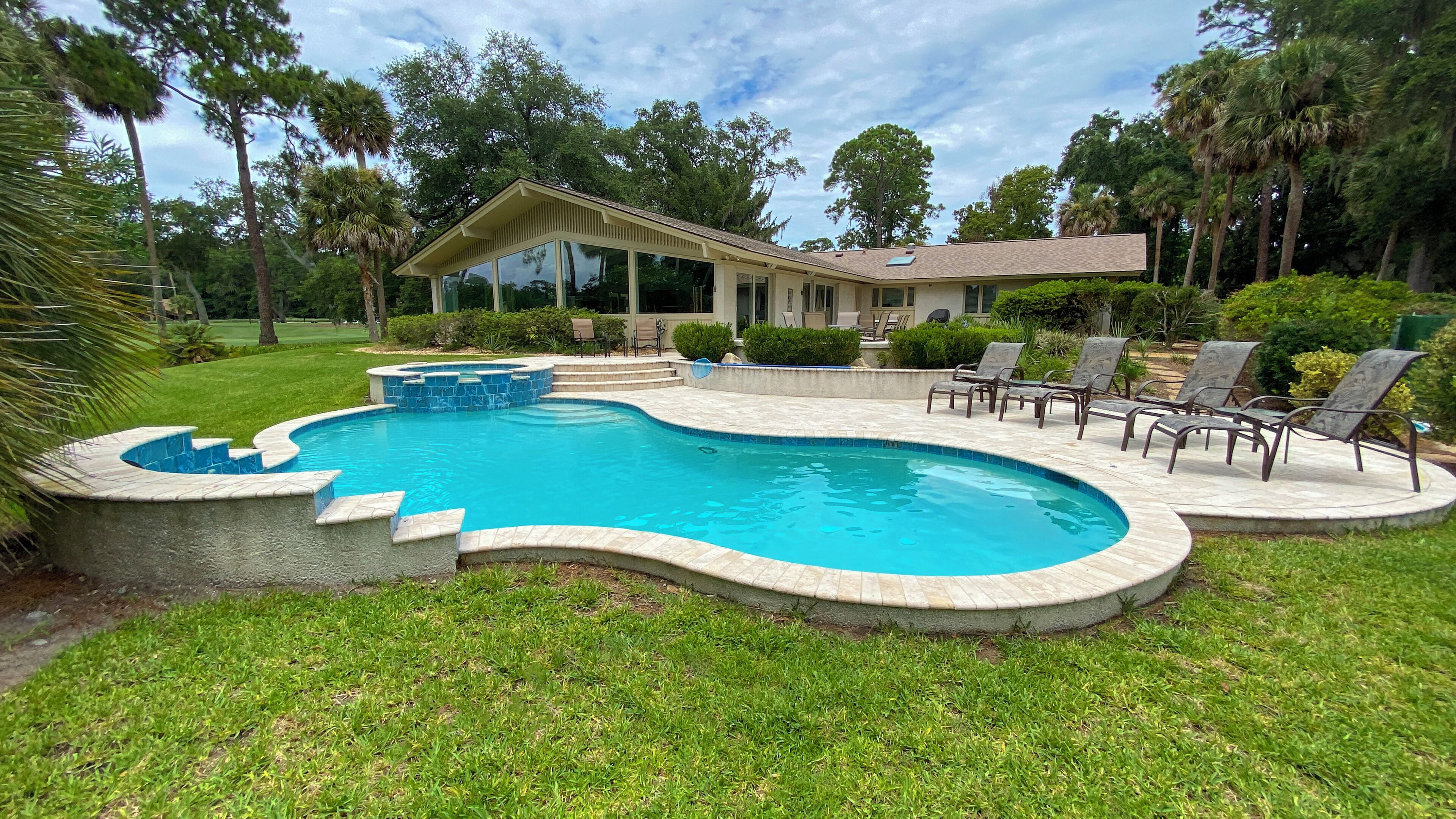 A swimming pool with a curved design, surrounded by a stone deck and a grassy area. There are lounge chairs and a covered patio area with a building in the background.