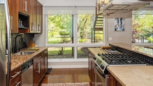 A modern kitchen with wooden cabinets, a stainless steel refrigerator, a gas stove, and a large window offering a view of a garden. - Juniper Presents: The Vineyard Loft - Cool Harbert Loft Near Beach with Fire Pit (Harbert)