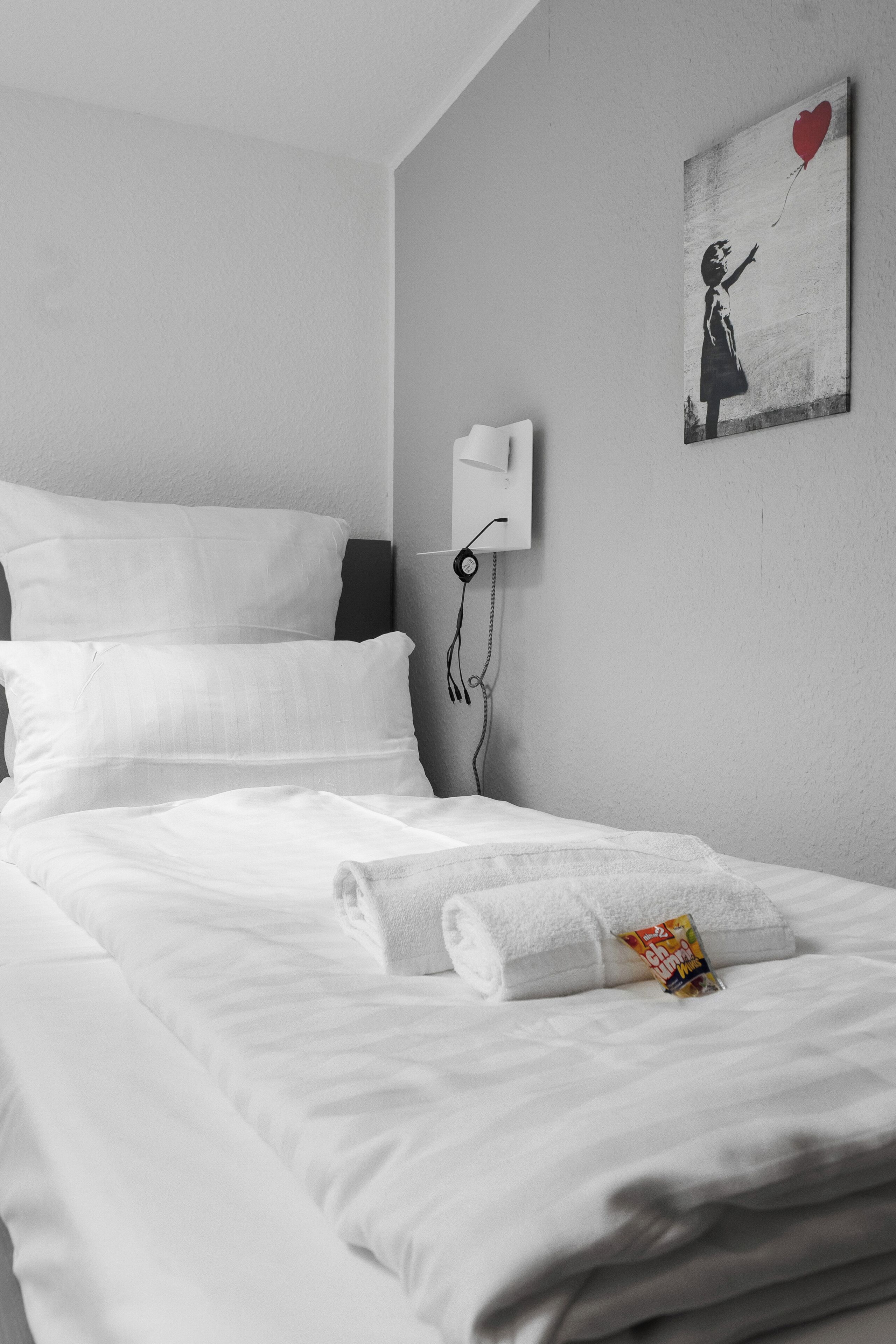 Un lit bien fait, avec du linge de lit blanc, une lampe fixée au mur et un tableau représentant une jeune fille tenant un ballon en forme de cœur.