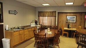 A hotel room with a wooden dining table, chairs, a kitchen area, and a framed picture on the wall.