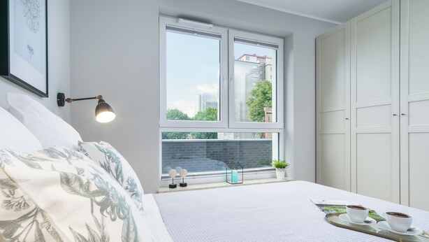 A bedroom with a bed, a window with a view of buildings, a bedside table with a lamp, and a tray with cups and saucers. - Wałowa - Garden View by Grand Apartments (Gdansk)