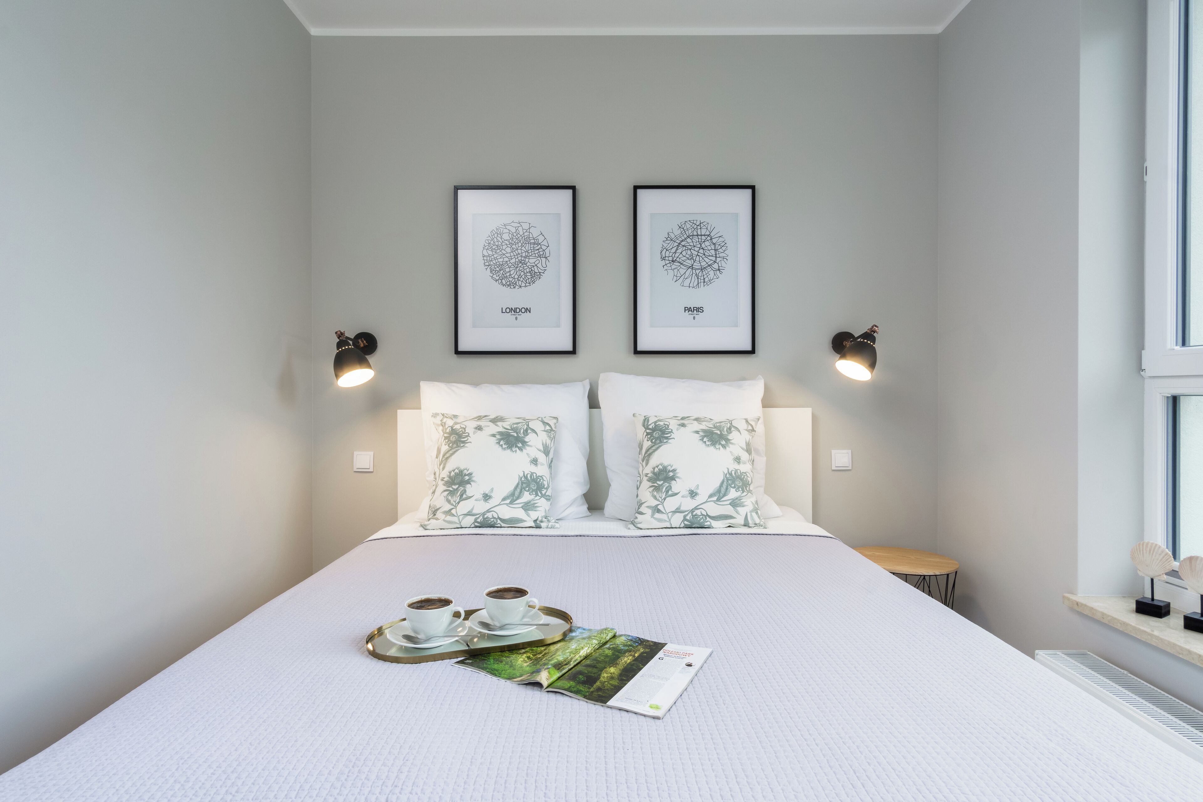Een net opgemaakt bed met een dienblad met koffiekopjes en een tijdschrift, twee aan de muur hangende kunstwerken met de tekst 'LONDON' en 'PARIS', en twee wandlampen.