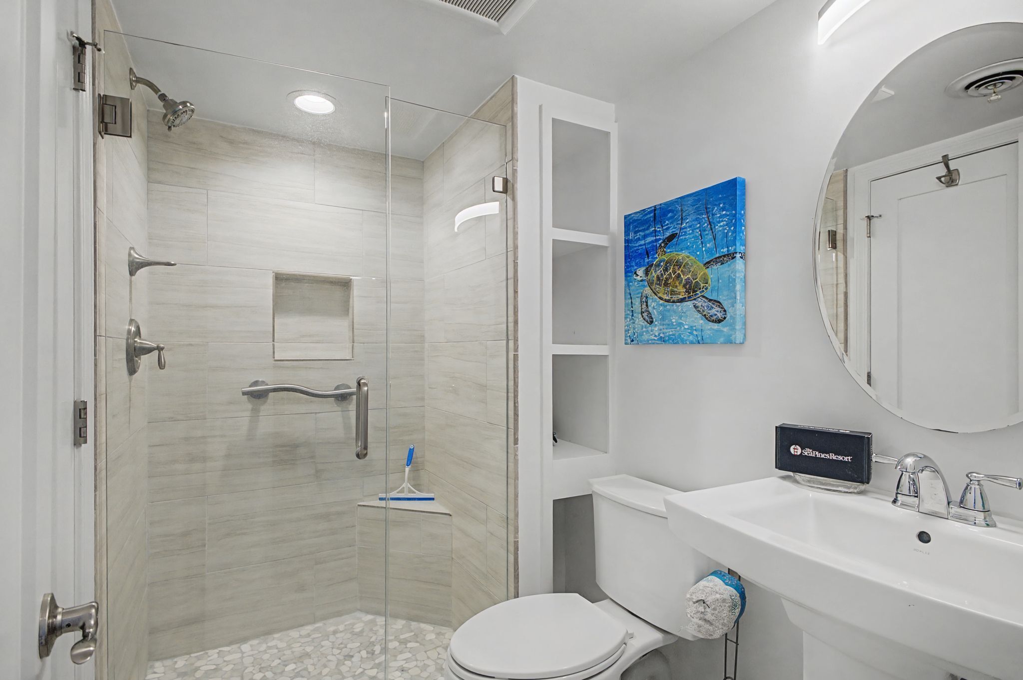 Un bany modern amb dutxa de vidre, vàter blanc, un lavabo amb mirall i un quadre de tortuga a la paret.