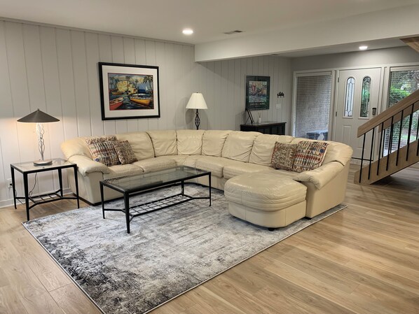 A living room with a beige sectional sofa, a glass coffee table, a patterned rug, a lamp, and a painting on the wall. - 459 Plantation Club Villas at The Sea Pines Resort (Hilton Head Island)