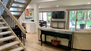 A spacious living area with a wooden staircase, a dining area, a sofa, a TV, and a large window offering a view of the outdoors. - 459 Plantation Club Villas at The Sea Pines Resort (Hilton Head Island)