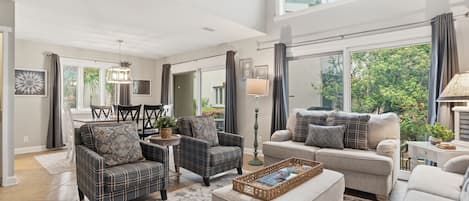 A spacious living room with a high ceiling, large windows, and a view of greenery outside.