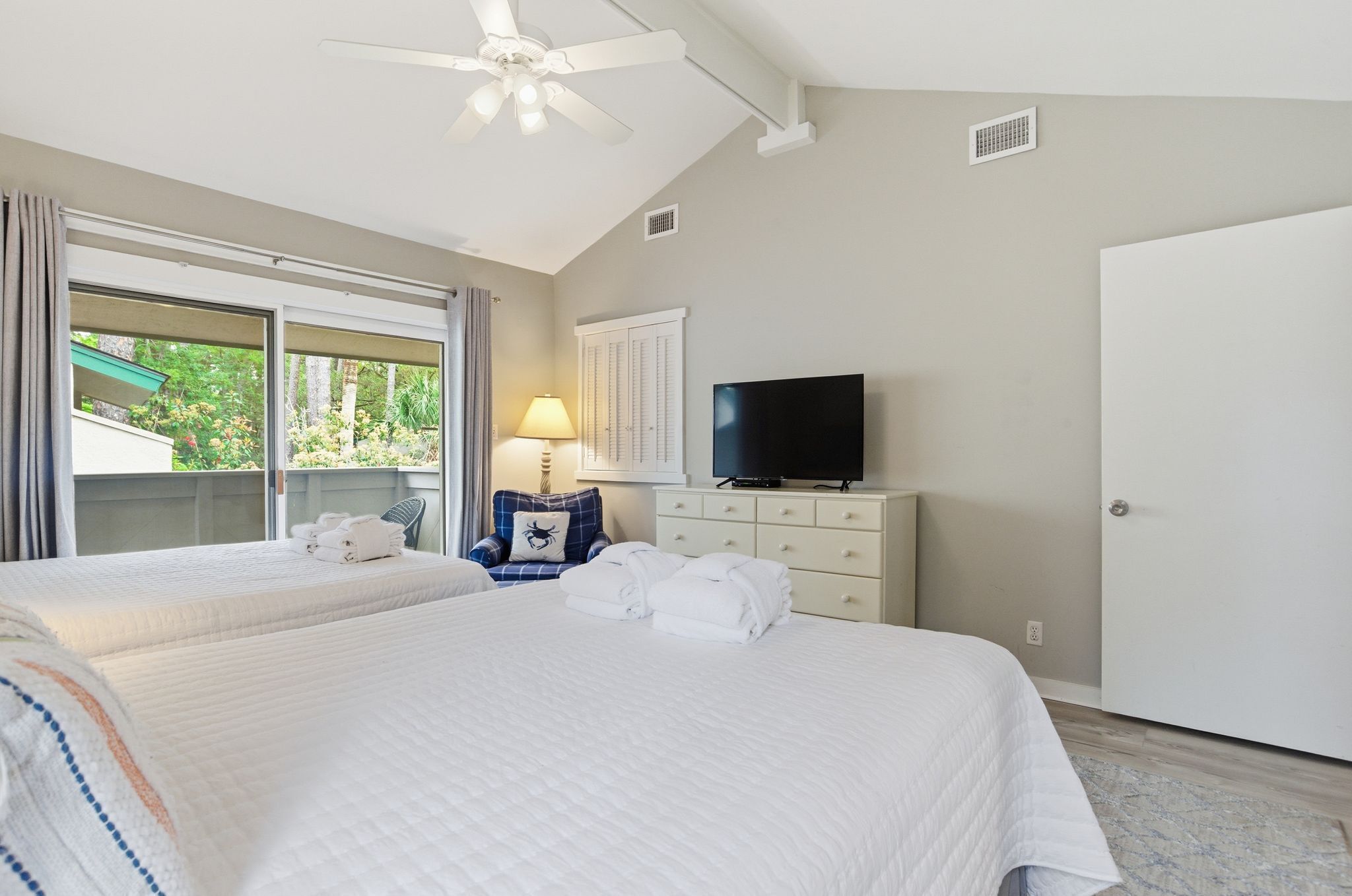 A hotel room with two beds, a TV, a ceiling fan, a dresser, and a view of greenery outside.