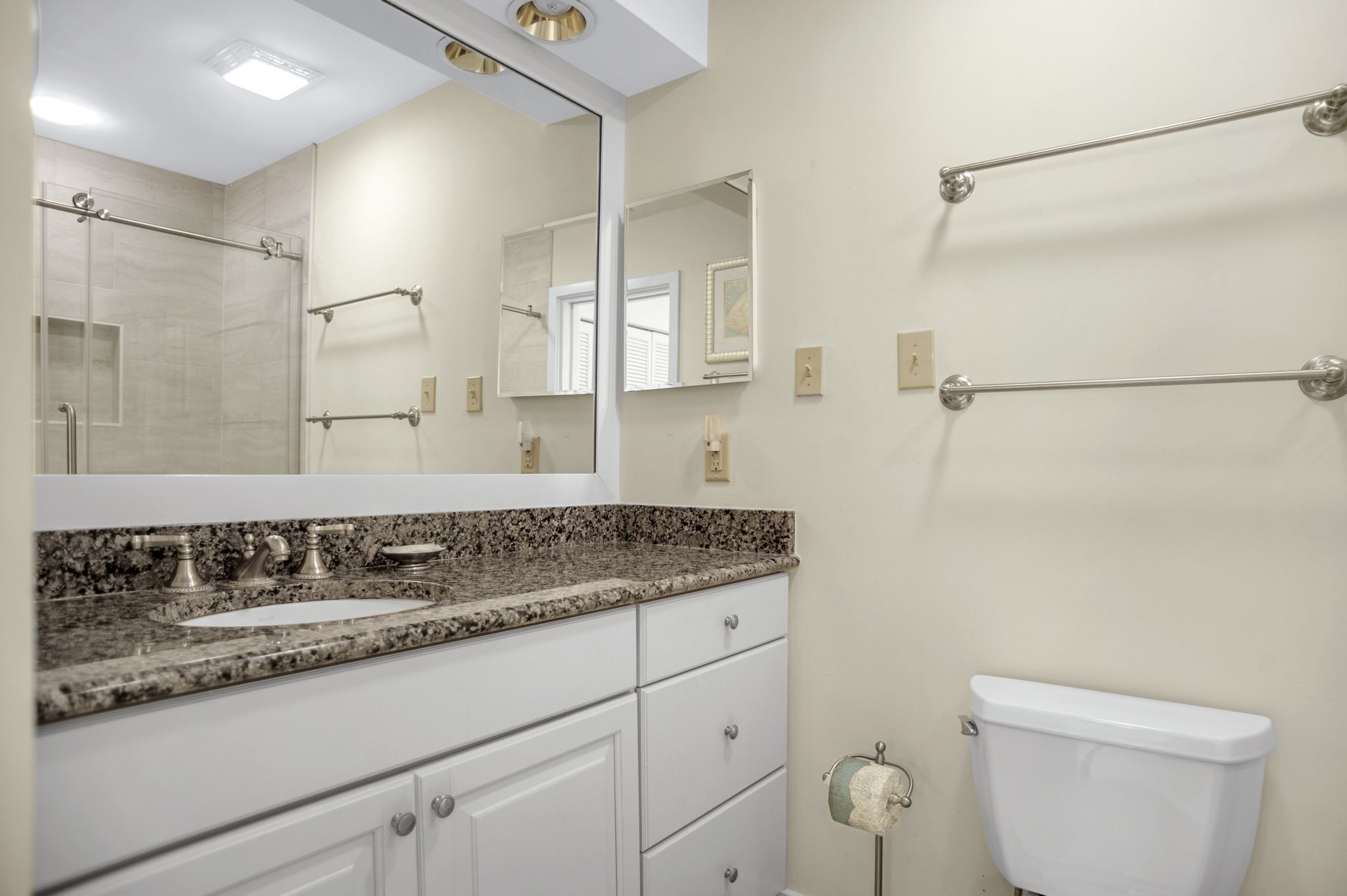 A bathroom with a granite countertop sink, white cabinets, a large mirror, a towel rack, and a toilet.