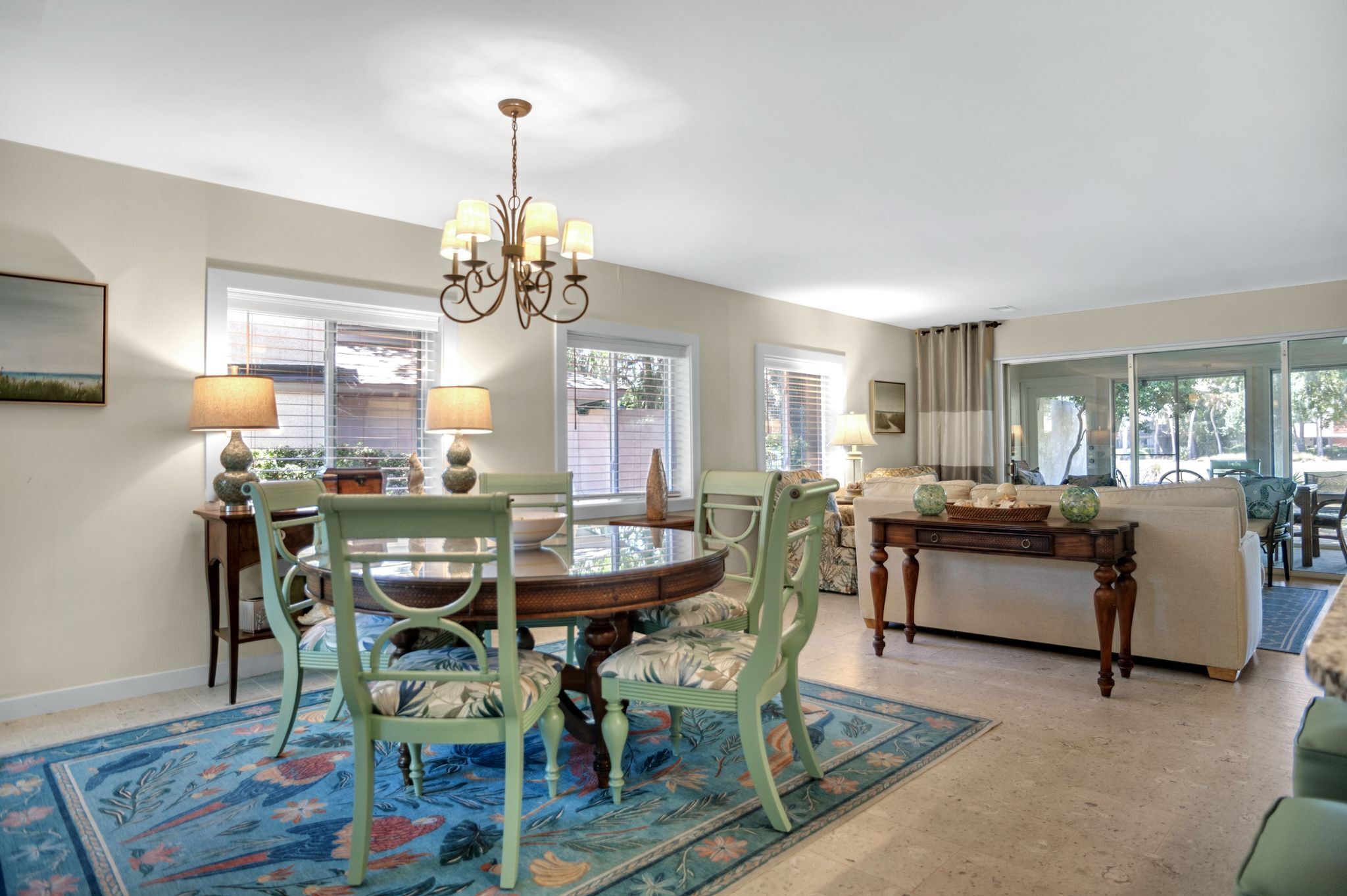 A dining area with a round table, chairs, and a chandelier. There are windows with blinds, a sofa, and a coffee table.