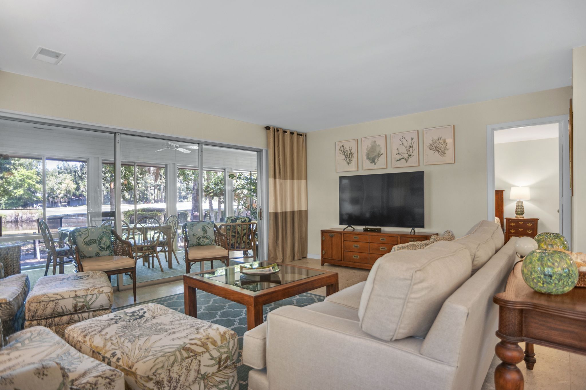 A spacious living room with a sofa, coffee table, and dining area with chairs. A flat-screen TV is mounted on the wall, and there are framed pictures above it.