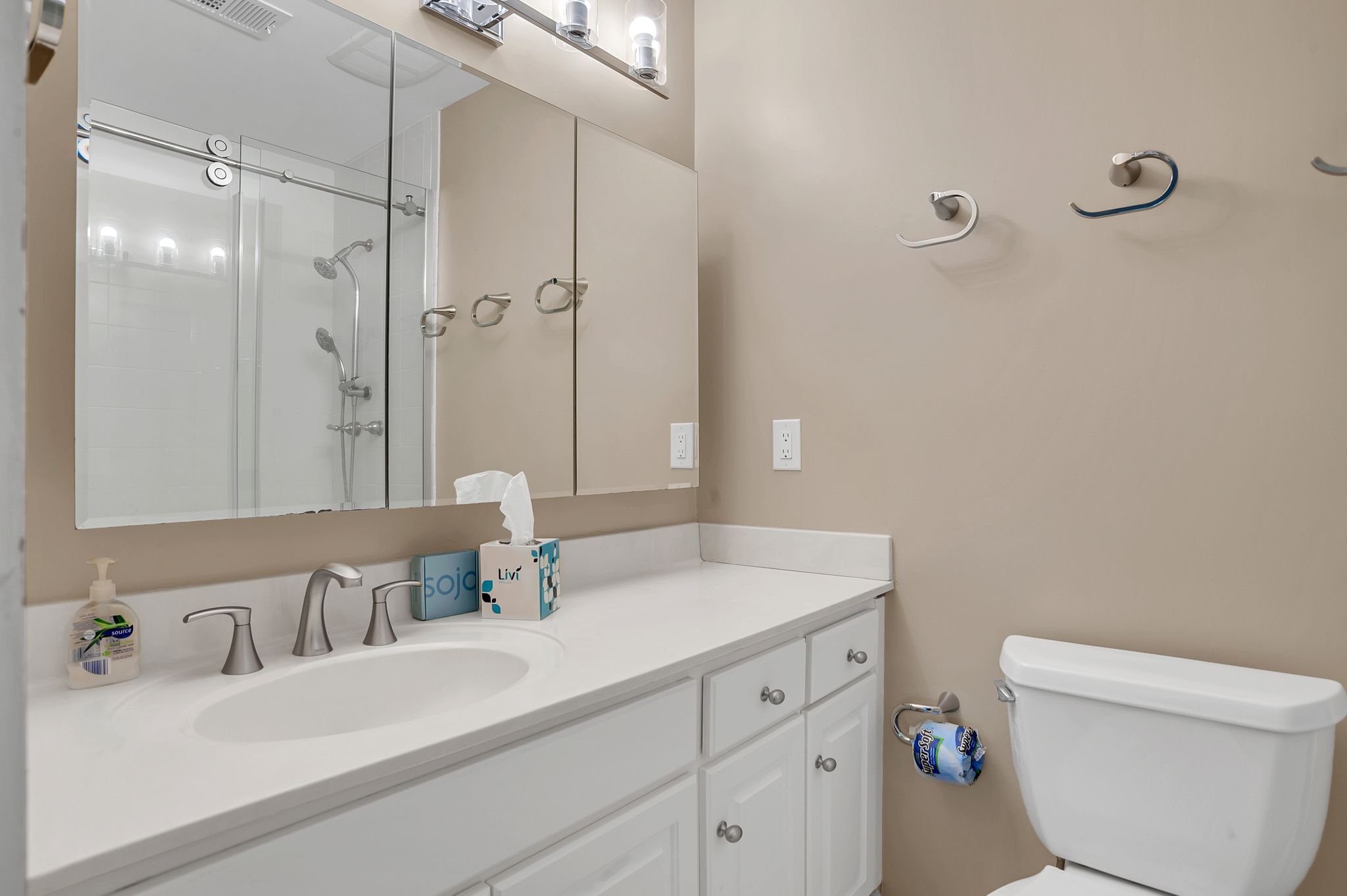 A bathroom with a white sink, mirror, and toilet. There are two towel racks on the wall, a soap dispenser, and a tissue box on the counter.