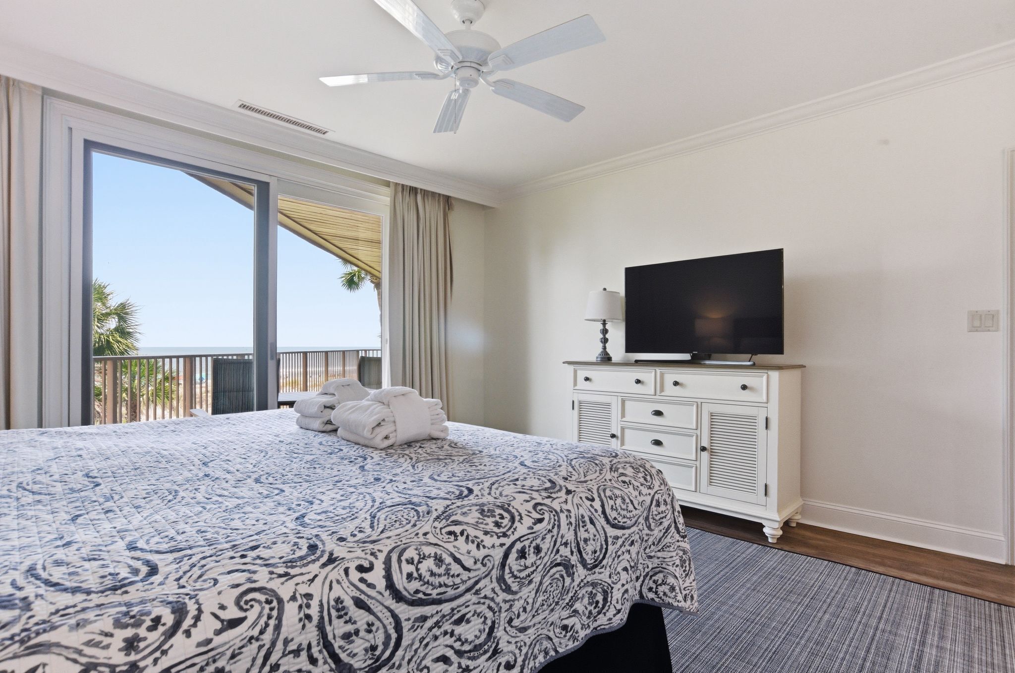 Ein Hotelzimmer mit einem großen Bett, einem Fernseher auf einer Kommode, einem Deckenventilator und einem Balkon mit Vorhängen.