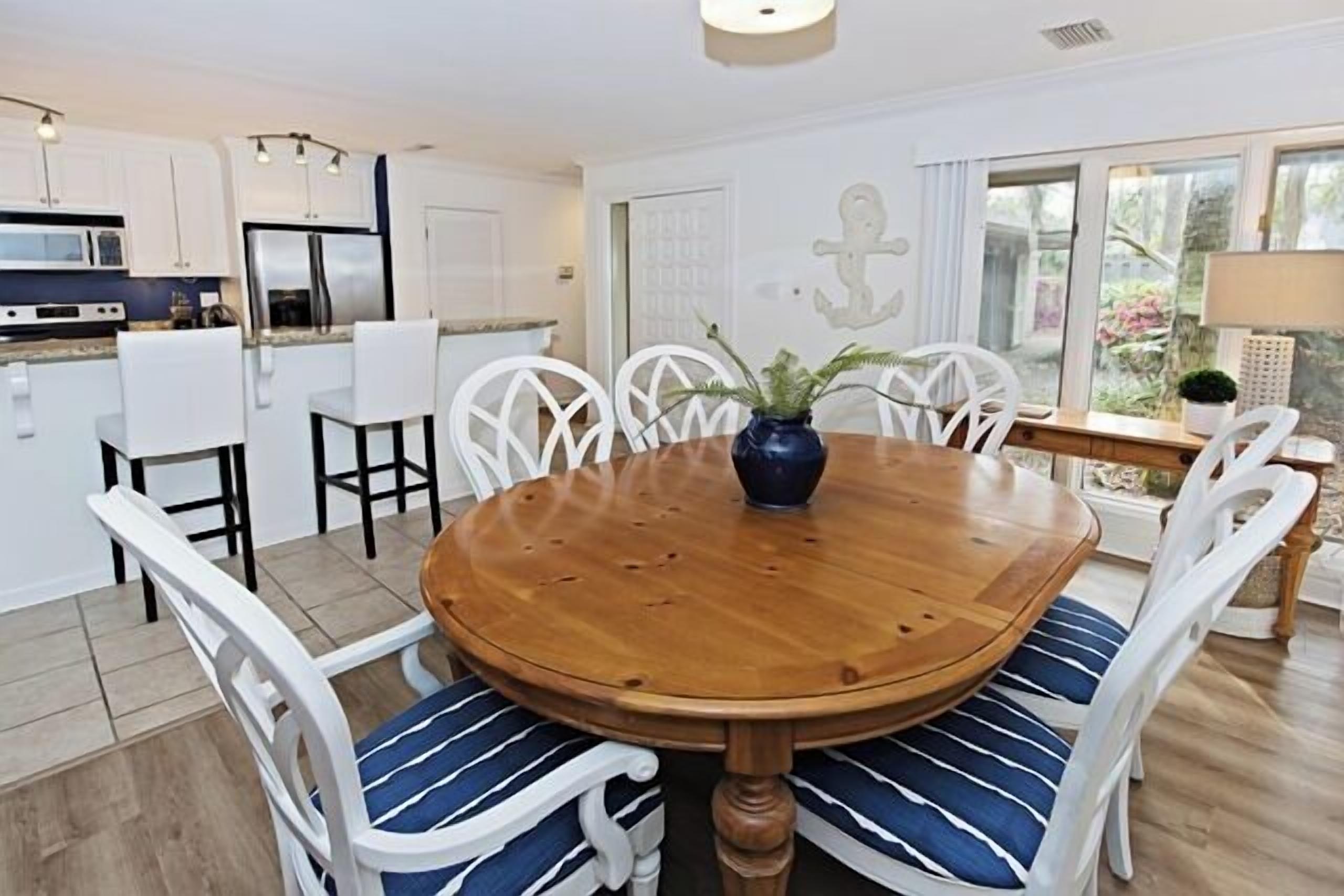 A dining area with a wooden table and white chairs, a kitchen in the background, and a view of greenery outside.