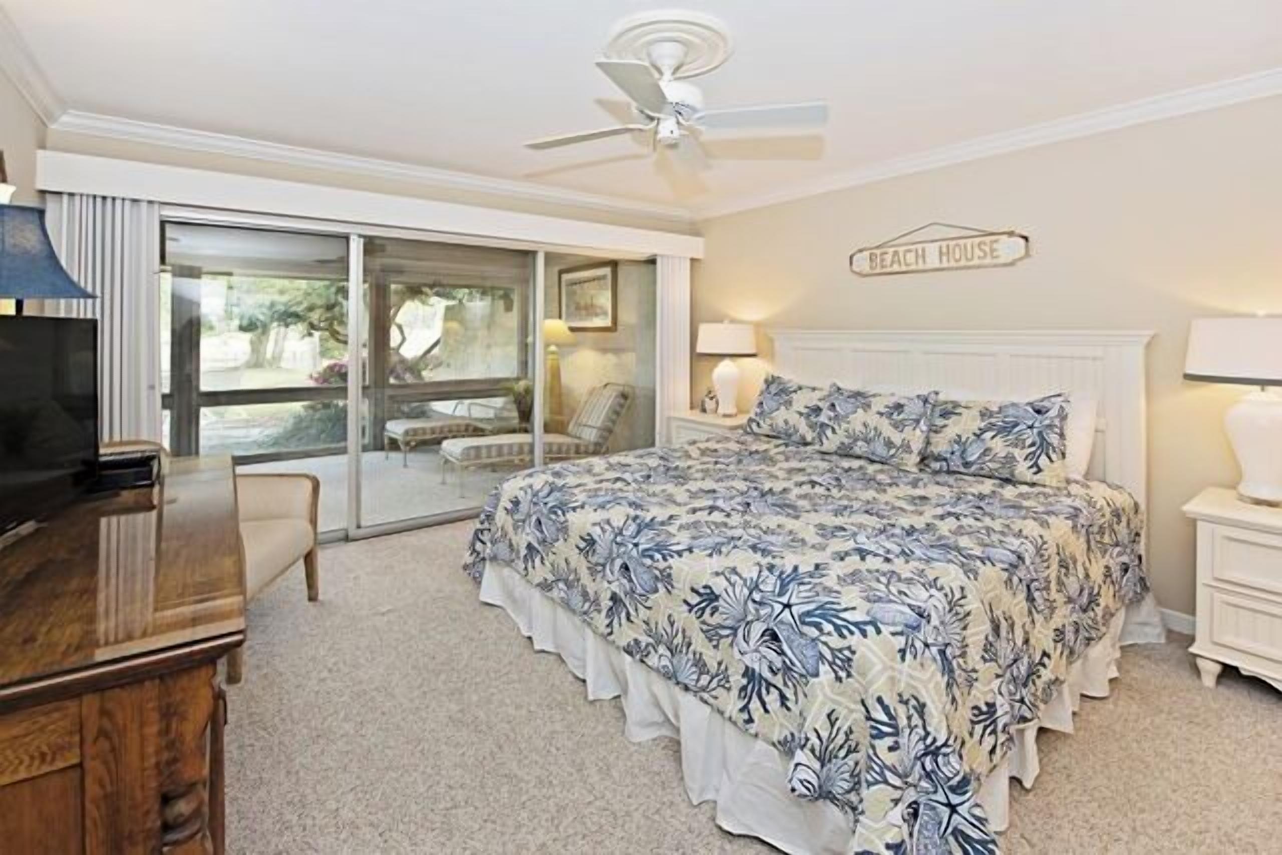 Une chambre avec un lit, une télévision, un ventilateur de plafond, une porte-fenêtre coulissante et une pancarte sur laquelle on peut lire « MAISON DE PLAGE ».