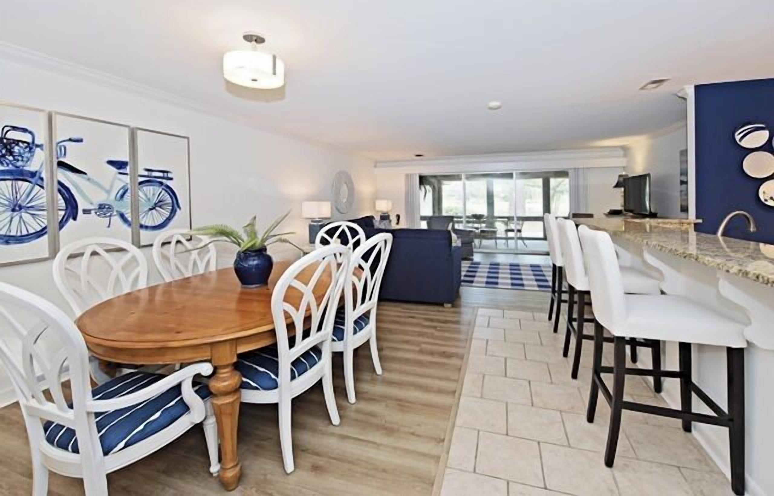A modern dining area with a wooden table and white chairs, a kitchen with a bar, and a view of the living area.