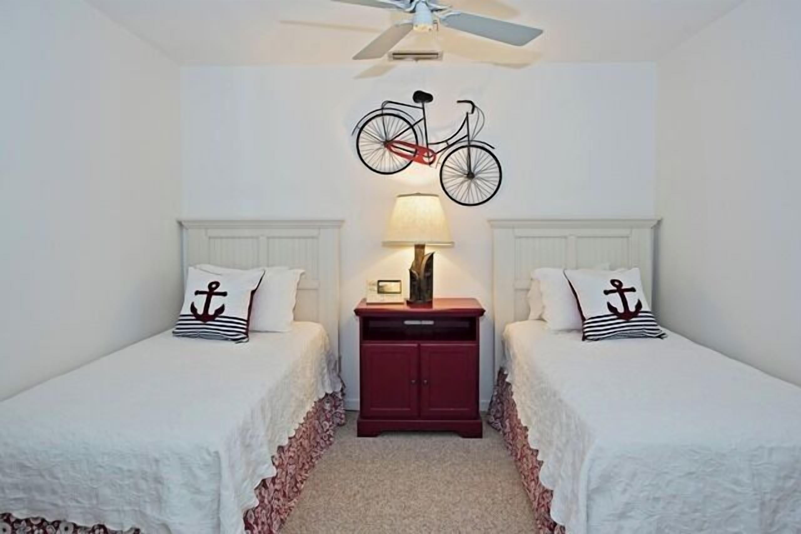A room with two beds, a red nightstand, a wall-mounted bicycle decoration, and a ceiling fan.
