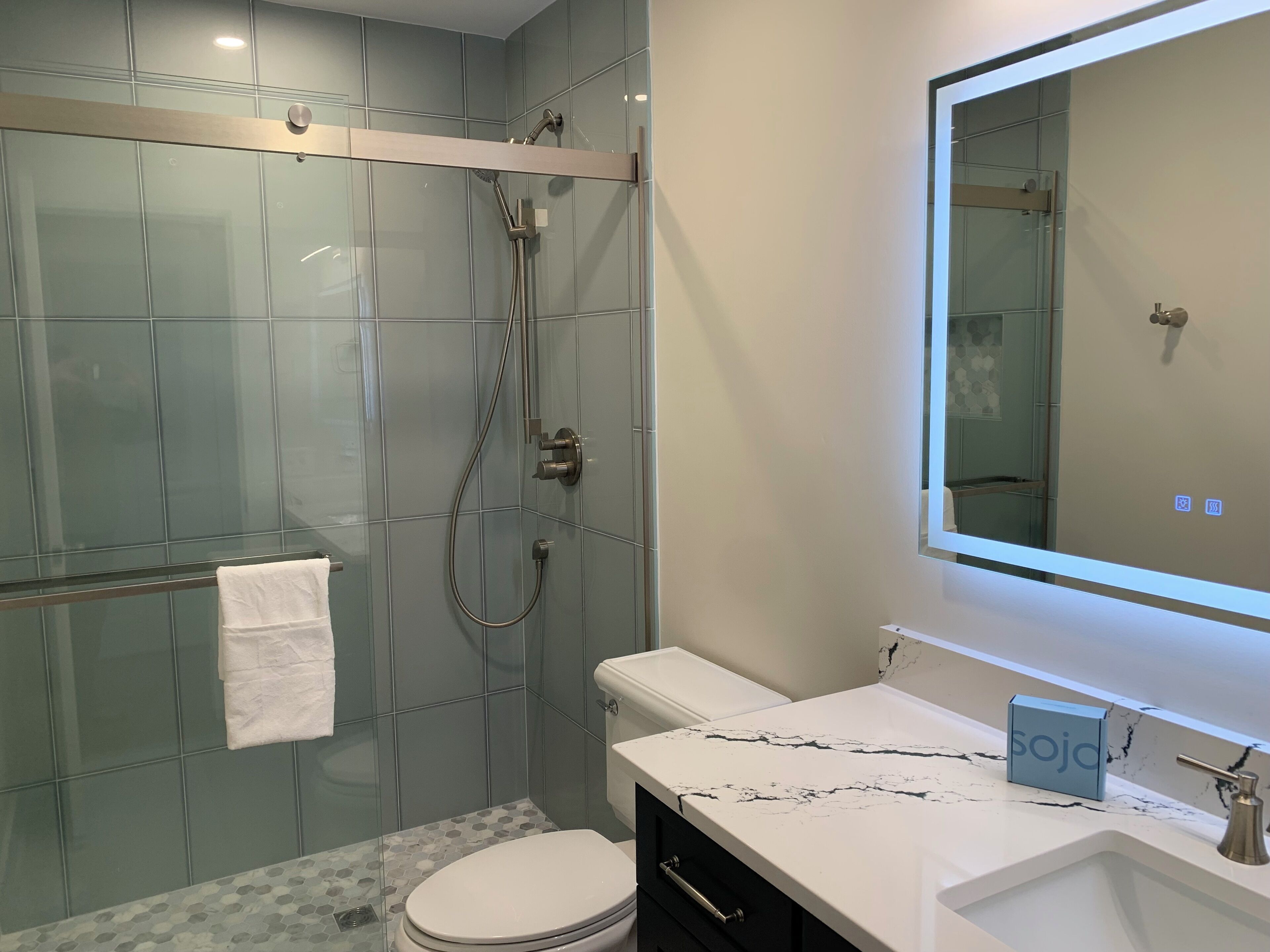 A modern bathroom with a glass shower enclosure, a white toilet, a marble countertop, and a large mirror.
