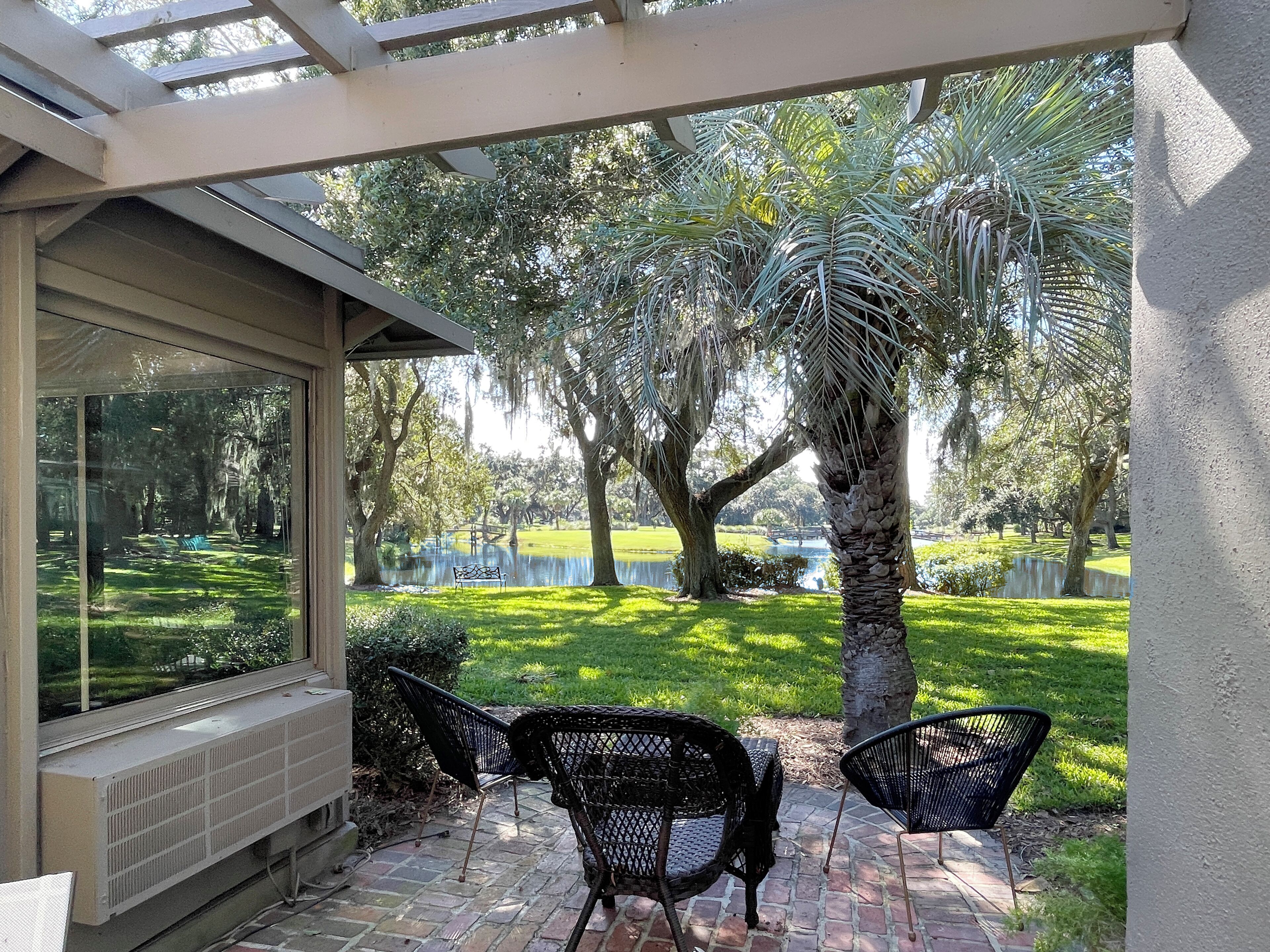 A patio with two black metal chairs and a table, overlooking a grassy area with trees and a body of water.
