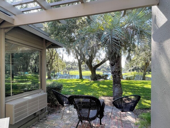 A patio with two black metal chairs and a table, overlooking a grassy area with trees and a body of water.
