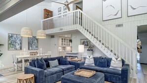 A spacious living area with a large blue sectional sofa, a small round stool, and a woven tray on an ottoman. A white staircase with a railing leads to an upper level. - 80 Fairway Lane at The Sea Pines Resort (Hilton Head Island)