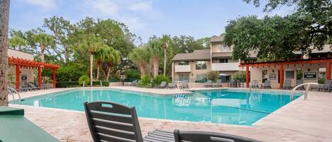 A swimming pool with lounge chairs and a covered seating area, surrounded by trees and buildings.