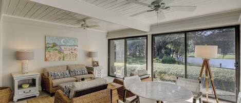 A spacious room with a ceiling fan, a large window with a view of a garden, wicker furniture, and a round table.