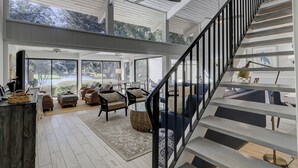 A spacious living area with a high ceiling, large windows, and a staircase with black railings. - 82 Fairway Lane at The Sea Pines Resort (Hilton Head Island)