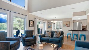 A spacious living area with a sofa, armchairs, and a glass coffee table. There is a kitchen area with a refrigerator and bar stools.
