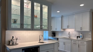 A modern kitchen with white cabinetry, a dishwasher, a coffee maker, and a glass-fronted cabinet displaying glassware.