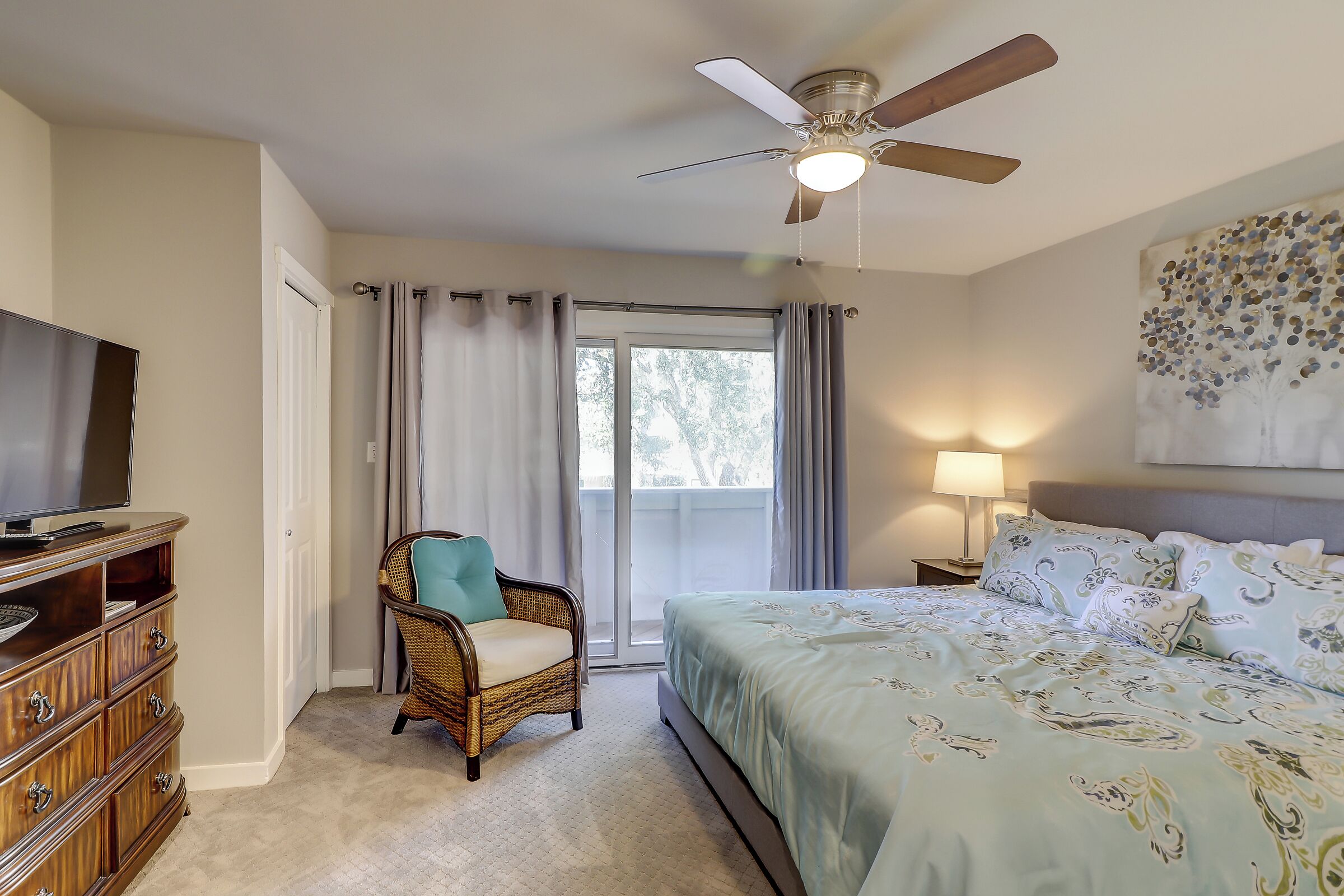 A bedroom with a bed, a television, a wicker chair, a ceiling fan, and a window with curtains.
