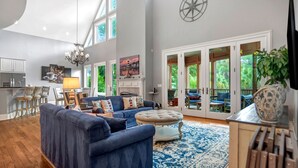 A spacious living room with a blue sofa, a round ottoman, and a patterned rug. There is a dining area with a chandelier, a kitchen with white cabinets, and a large window overlooking greenery. - 64 Lawton Road at The Sea Pines Resort (Hilton Head Island)