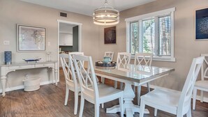 Un coin repas avec une table en verre, des chaises blanches, un luminaire suspendu et une fenêtre avec des stores.