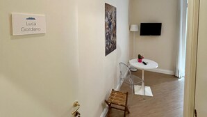 A hallway with a white wall, a door with a keychain, a small wooden chair, a table with a lamp, and a TV mounted on the wall. - de Ribera rooms (Naples)