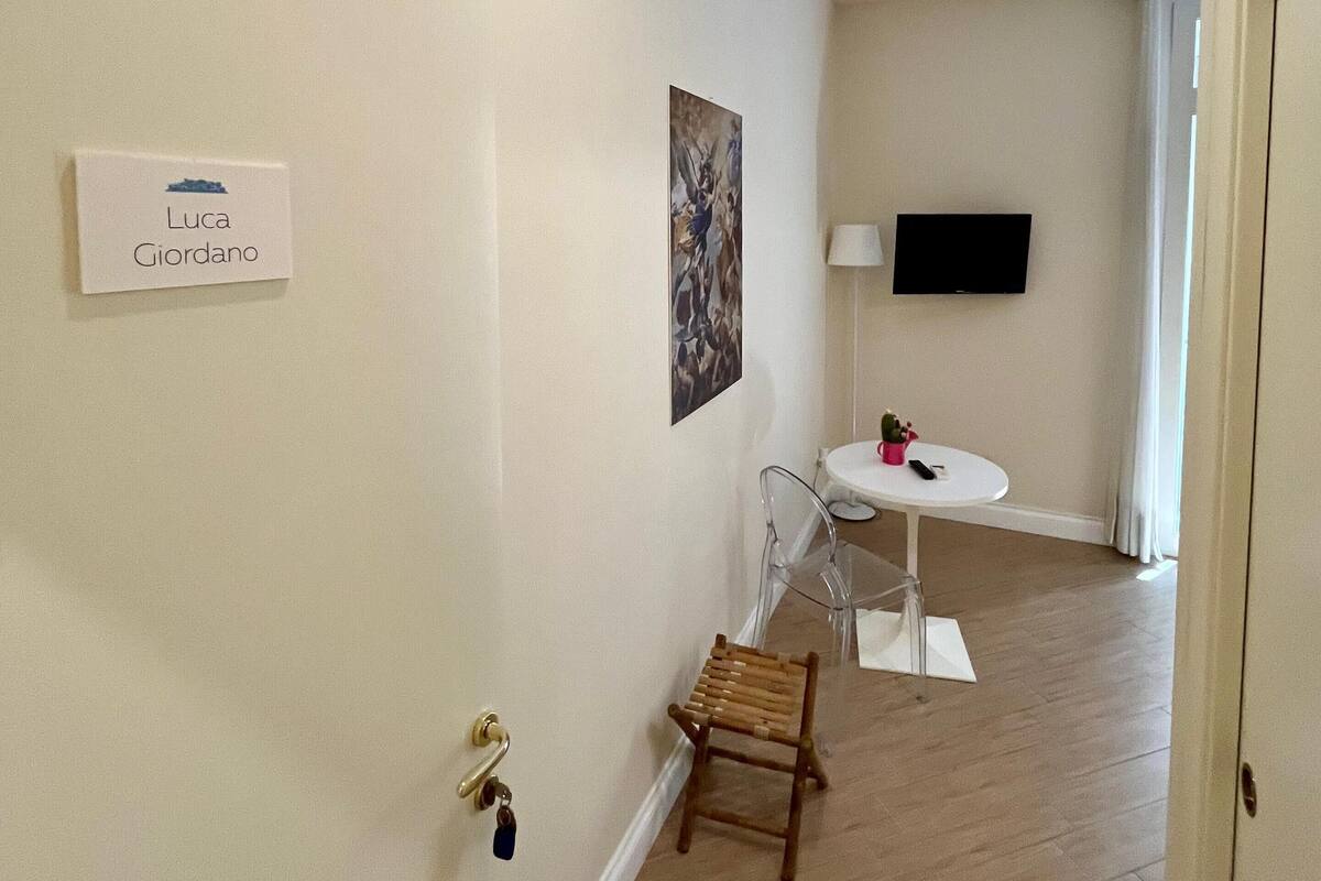 A hallway with a white wall, a door with a keychain, a small wooden chair, a table with a lamp, and a TV mounted on the wall.