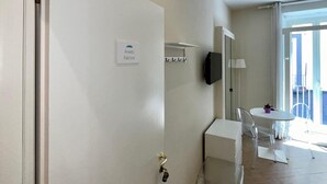 A hotel room with a door labeled "Anieleo Falcone," a small white table with chairs, a TV mounted on the wall, and a window with a view of the outside. - de Ribera rooms (Naples)